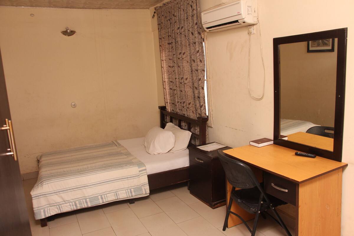 A small, single-bed room with a wooden desk, a chair, a mirror, and a bedside table.
