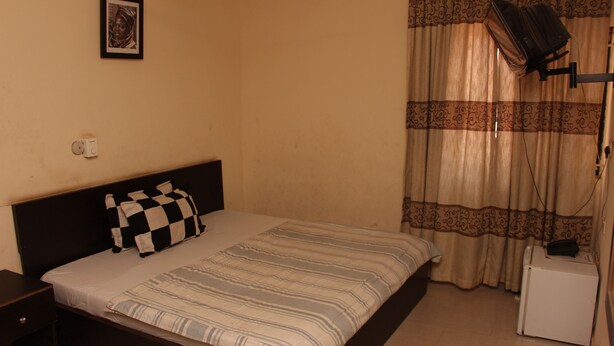 A bedroom with a bed, a nightstand, a small table, a wall-mounted TV, and a curtain. - Sapphire Guest House (Abuja)