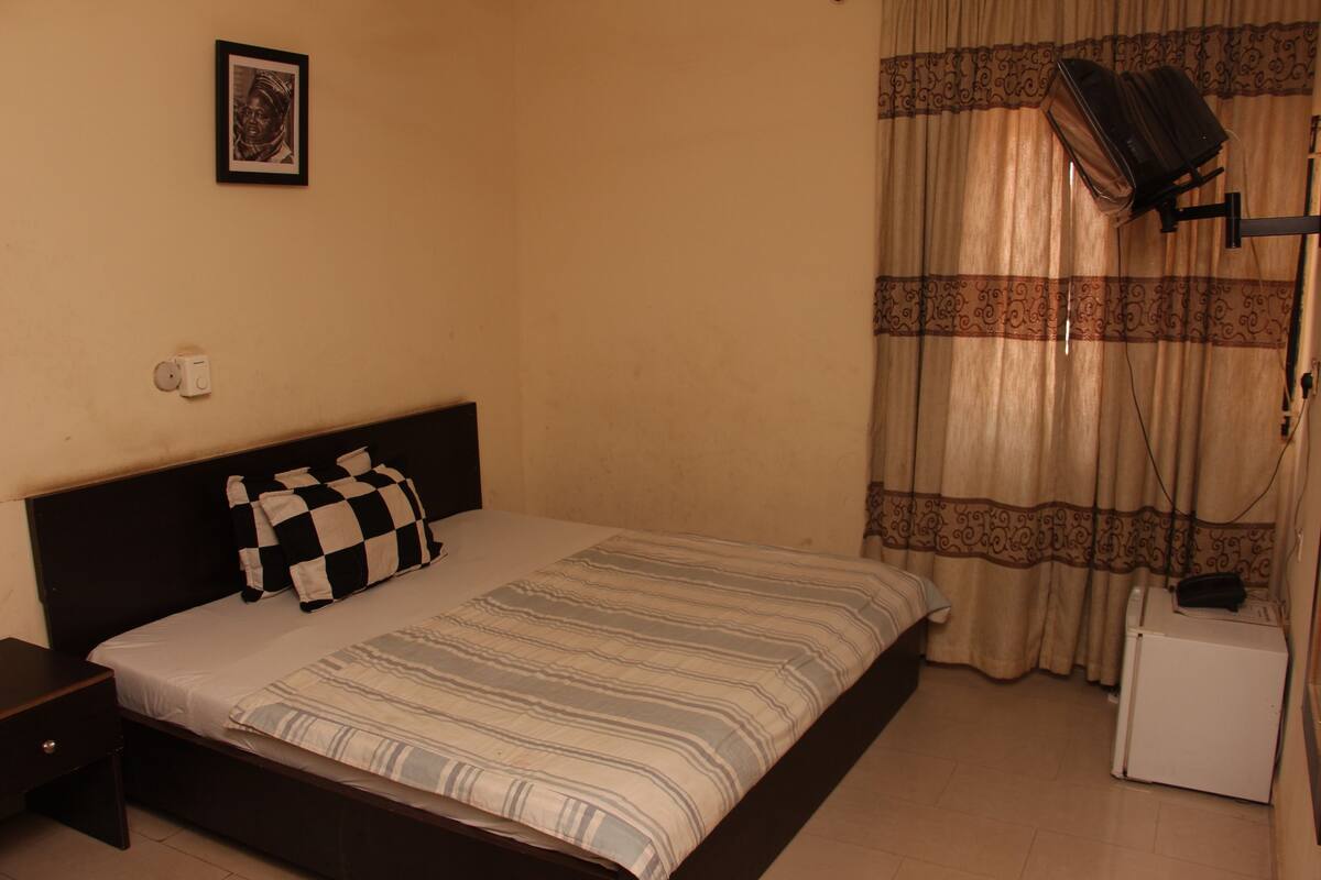 A bedroom with a bed, a nightstand, a small table, a wall-mounted TV, and a curtain.