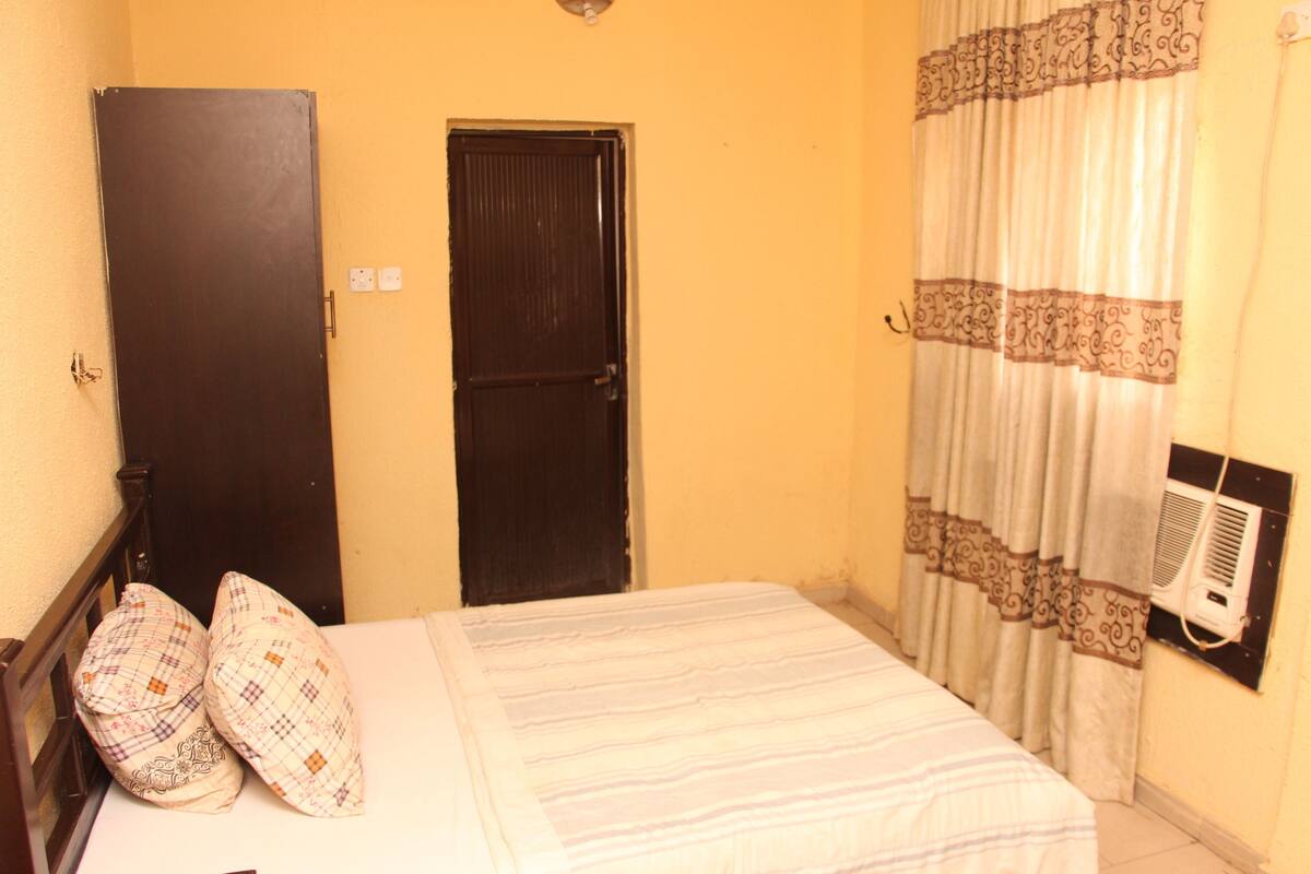 A bedroom with a bed, two pillows, a wardrobe, a door, and a wall-mounted air conditioner.