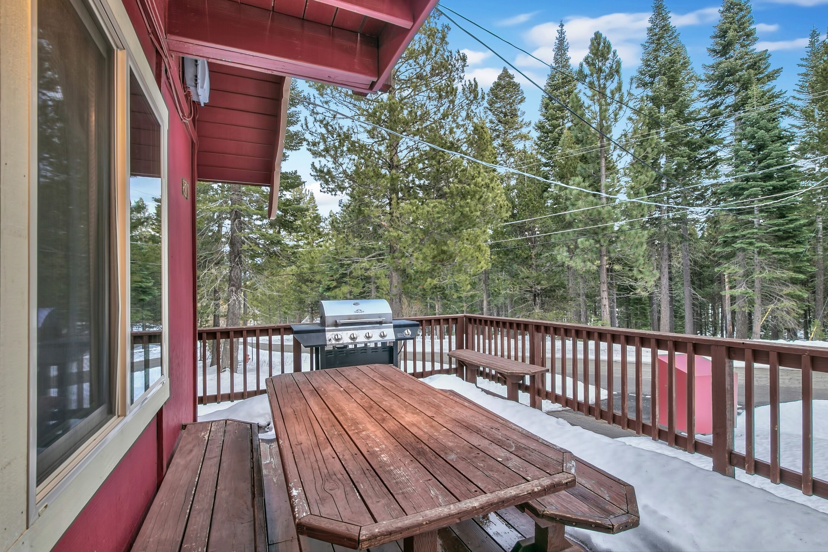 A wooden deck with a barbecue grill, a picnic table, and a view of a snowy landscape with tall trees.