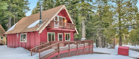 A red cabin with a balcony, surrounded by snow and tall trees.
