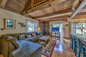 A cozy living room with a stone fireplace, wooden ceiling beams, a sofa, and a dining area with chairs.