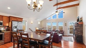 Una cocina amplia con armarios de madera, una gran mesa de comedor y un candelabro. Cuenta con una chimenea de piedra y vistas al exterior a través de ventanas grandes.