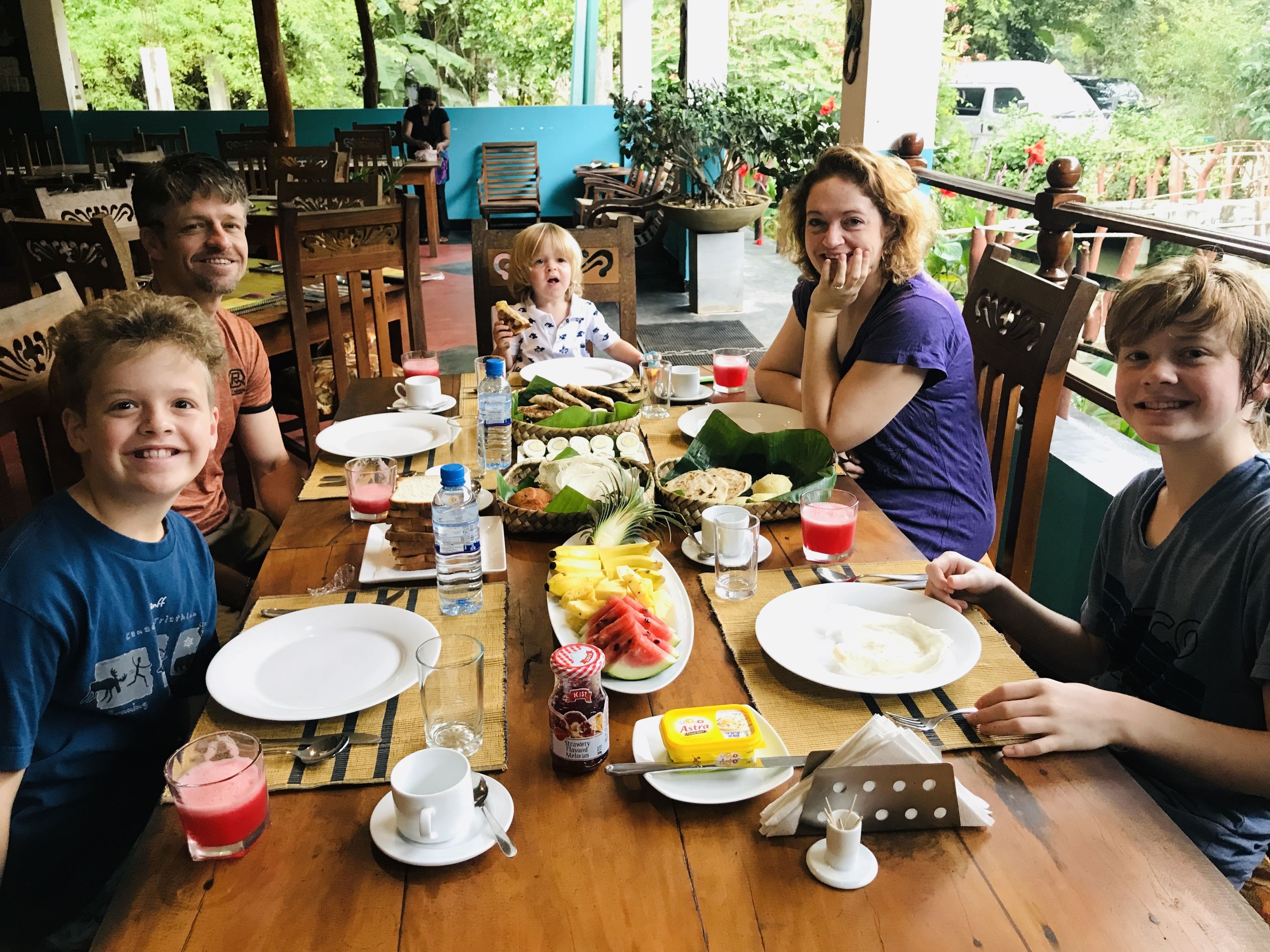 A family dining at a wooden table with various dishes, drinks, and a view of greenery outside.