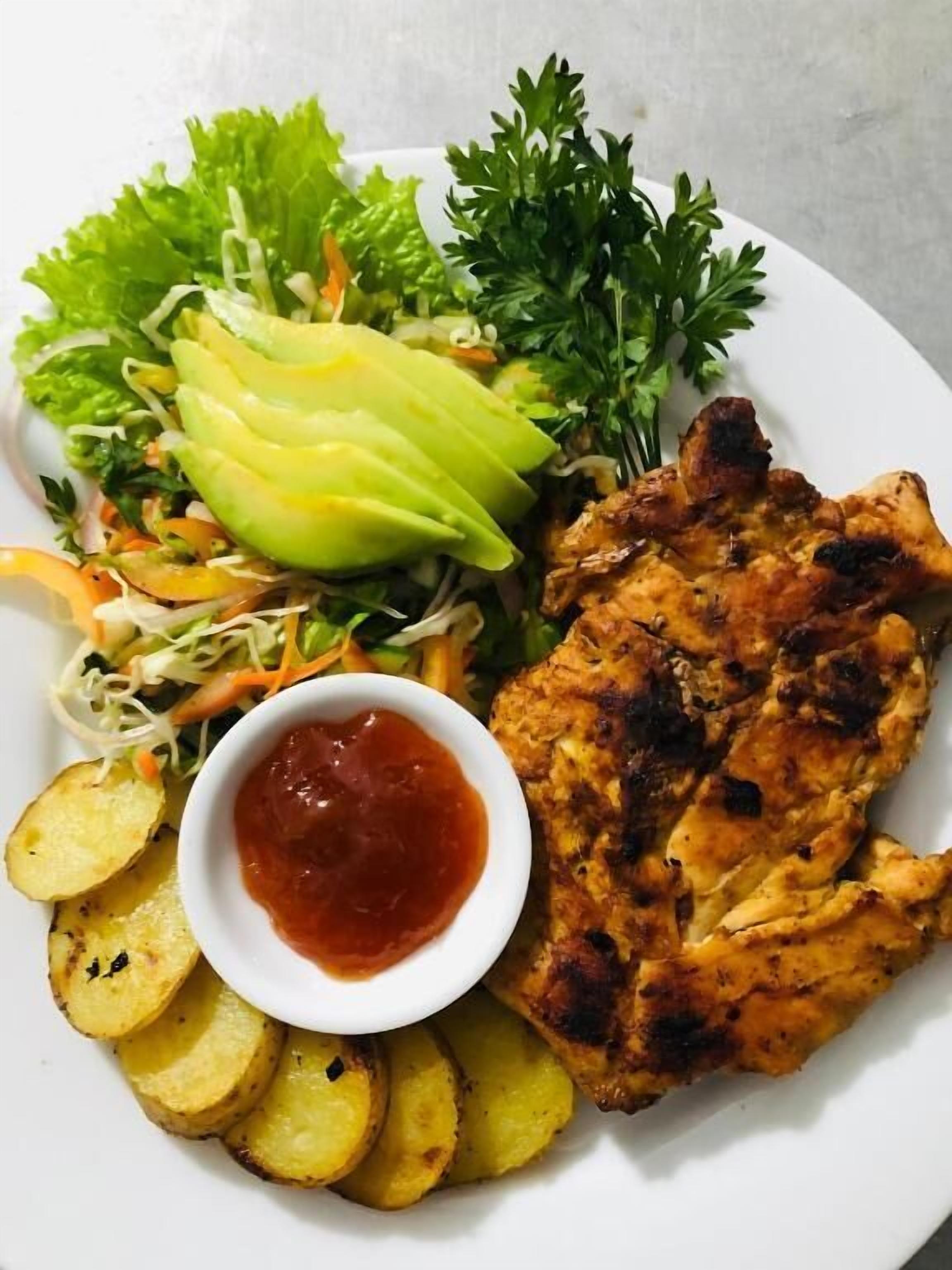 A plate with grilled chicken, a side salad, roasted potatoes, and a small bowl of sauce.