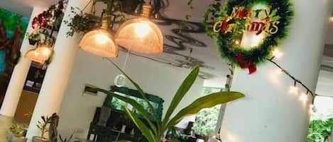 Un espacio interior decorado con luces colgantes, un adorno navideño que dice 'Merry Christmas' y plantas en macetas.