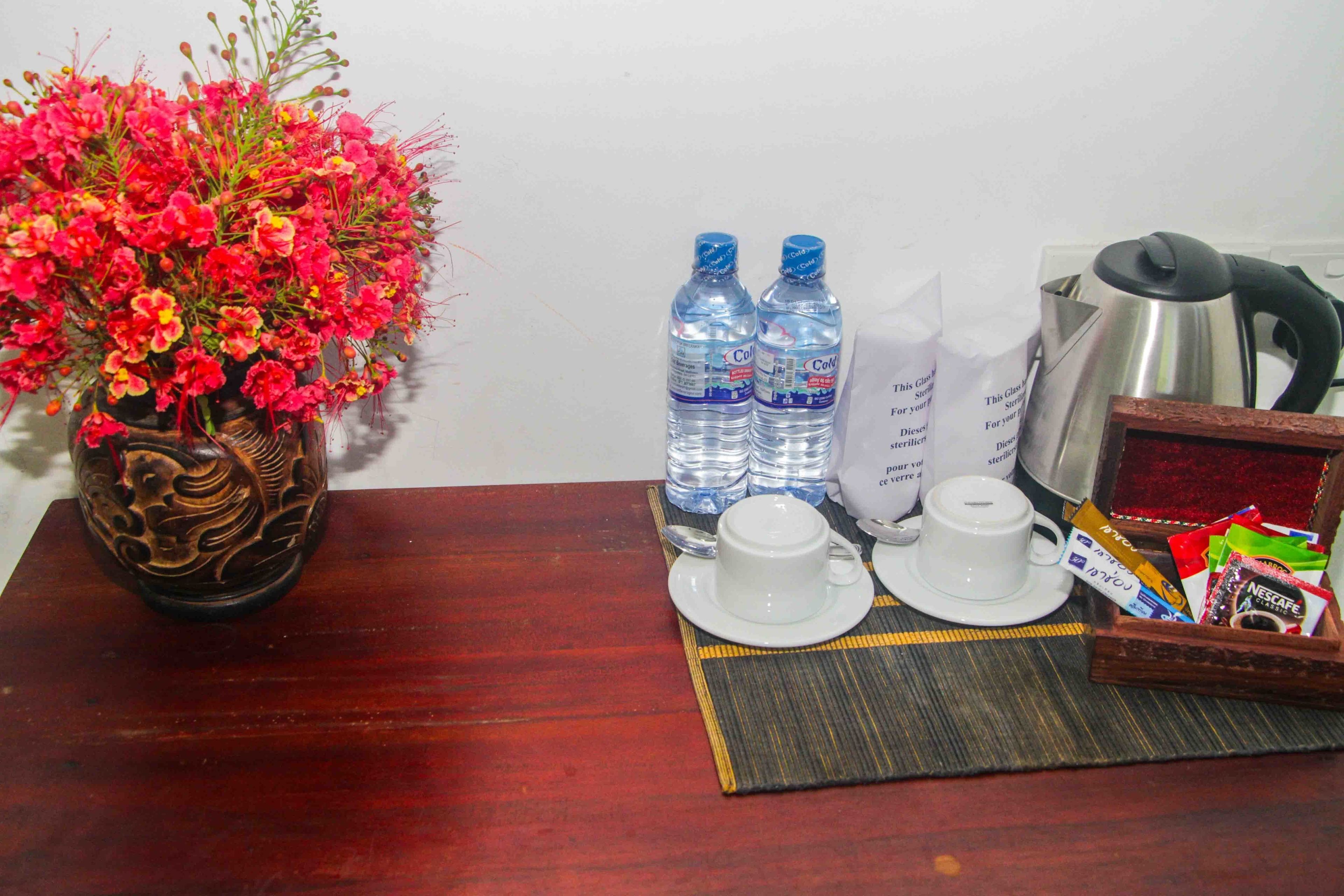 Une table en bois sur laquelle se trouvent un vase de fleurs, deux bouteilles d’eau, une cafetière, une tasse, une soucoupe et un plateau contenant des sachets de café.