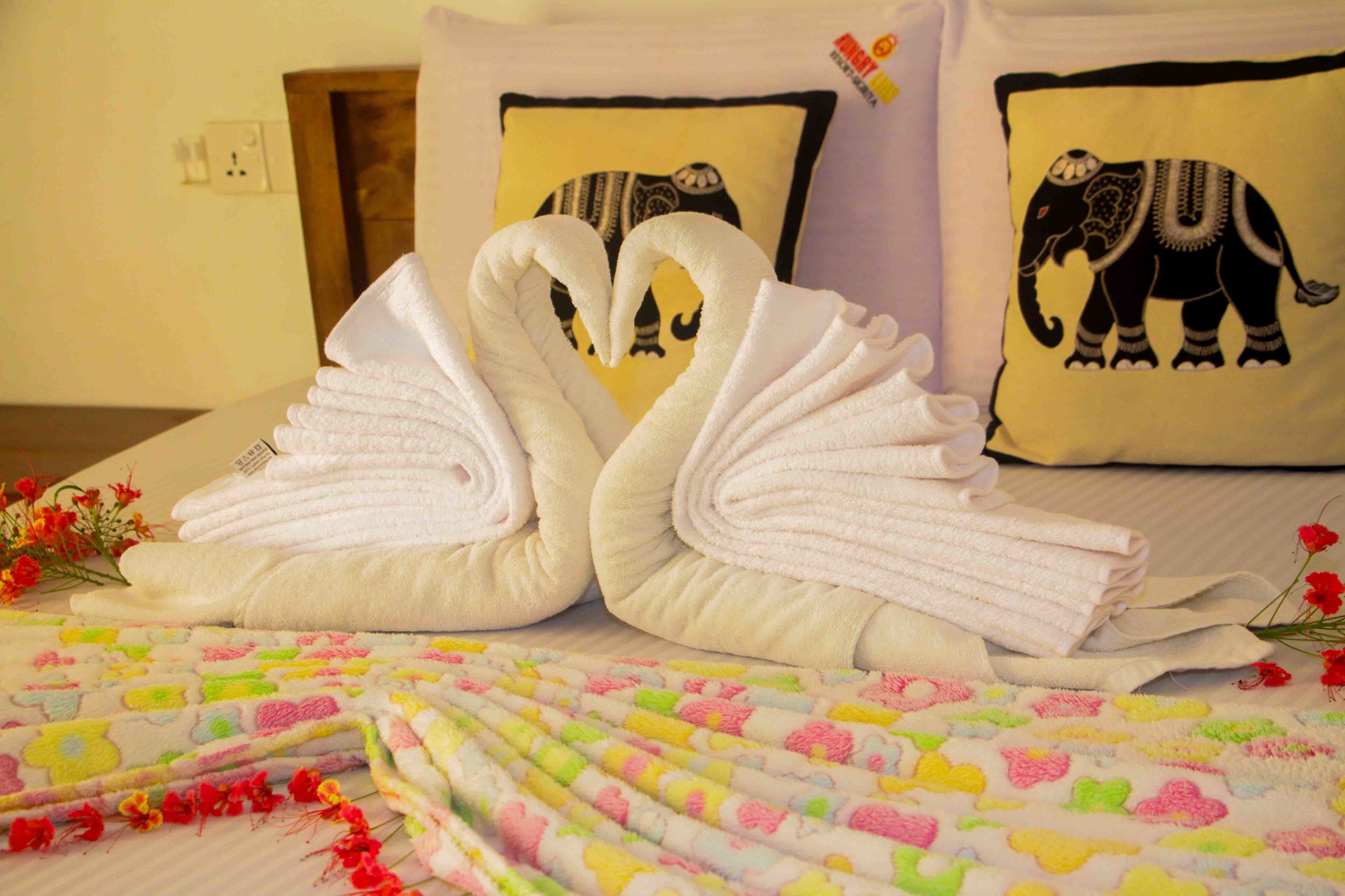 A neatly made bed with two towels arranged in the shape of swans, a colorful blanket, and decorative pillows with elephant designs.