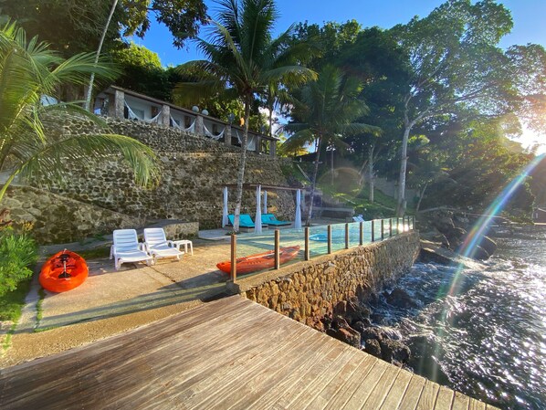 Una terraza de madera con sillones de descanso y un kayak, un muro de piedra y un área de piscina con vista al océano.