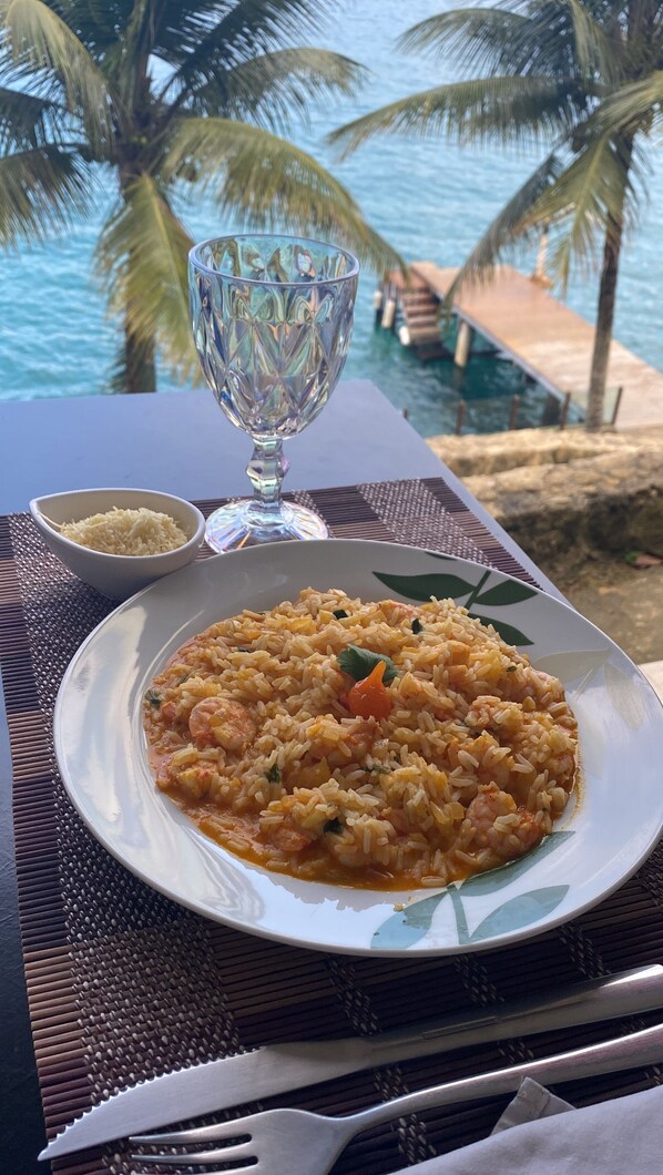 A plate of rice with vegetables and a glass of water on a table with a view of the ocean and palm trees. - Pousada Serra e Mar (Angra dos Reis)