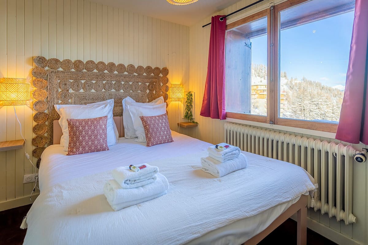 Un lit bien fait, avec du linge de lit blanc et des coussins à motifs roses, une tête de lit en osier et une vue sur un paysage enneigé par une fenêtre ornée de rideaux roses.