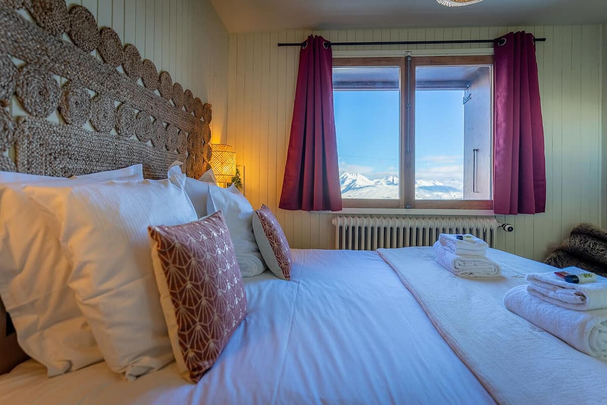 Une chambre avec un grand lit, une tête de lit en bois, une fenêtre avec des rideaux, un radiateur et une vue sur des montagnes enneigées.