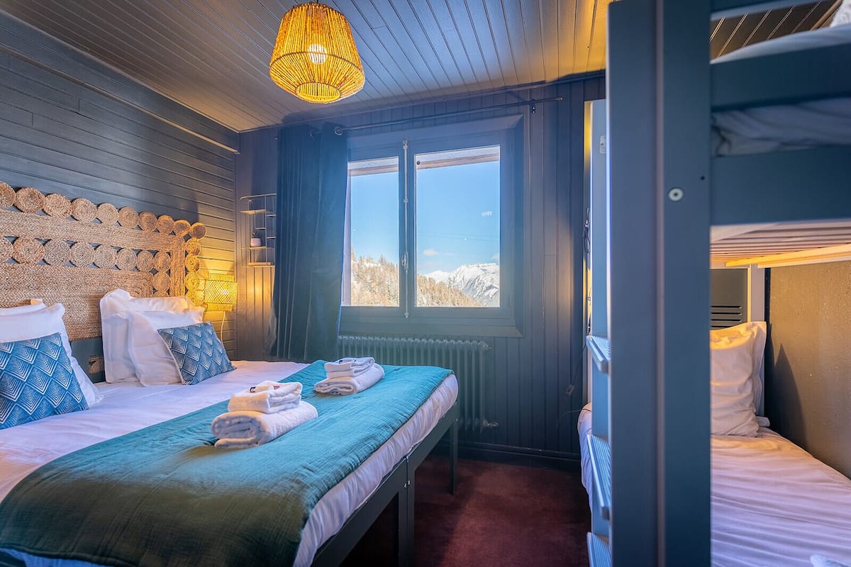 Une chambre accueillante avec un lit superposé, une grande fenêtre donnant sur les montagnes, une tête de lit en osier et un luminaire à la lumière chaleureuse.