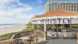 A beachside restaurant with outdoor seating, a view of the ocean, and a building in the background. - Royale Palms 2307 (Myrtle Beach)