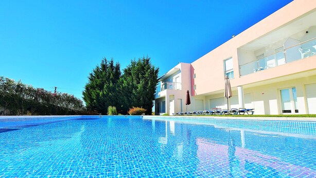Una piscina azul cristalina con fondo de azulejos, rodeada por un edificio moderno con balcones y un cielo despejado.