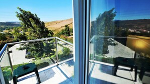 Un balcón con baranda de cristal, una silla verde y vistas a un paisaje montañoso con árboles y una carretera.