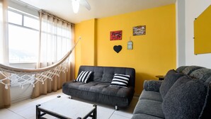 Un salon avec un hamac, un canapé gris, une table basse et un mur jaune orné d’objets décoratifs.