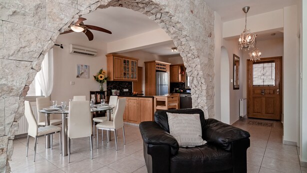 A spacious living area with a stone archway, a dining table with chairs, a black leather sofa, and a kitchen in the background. - Villa Puccini (Pegeia)