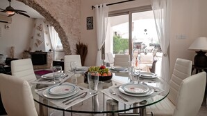 A dining area with a glass table set for four, surrounded by white chairs. A stone wall and a ceiling fan are visible in the background. - Villa Puccini (Pegeia)