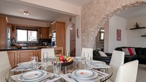 A modern kitchen with wooden cabinets and a stone archway leading to a living area with a black sofa and a glass dining table set for four. - Villa Puccini (Pegeia)