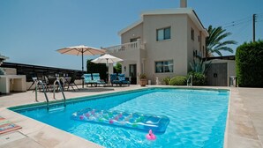 A swimming pool with lounge chairs and umbrellas, a building with balconies, and a palm tree. - Villa Puccini (Pegeia)