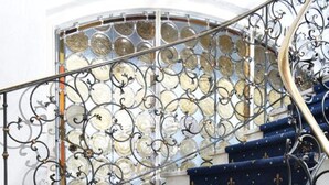 Eine Treppe mit blauem Teppich und goldenen Lilienmotiven, die zu einem Geländer aus Glas führt.