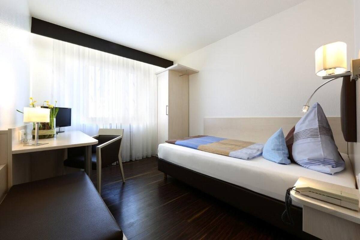 A hotel room with a bed, bedside table, lamp, desk, chair, and curtains.