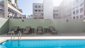 Une piscine entourée de chaises longues et dotée d’un escalier, le tout bordé d’un mur carrelé et avec des bâtiments en arrière-plan.
