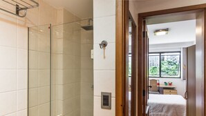 Une salle de bain dotée d’une douche à parois de verre, d’un mur recouvert de carreaux blancs et d’une porte en bois donnant sur une chambre à coucher avec un lit et une fenêtre.