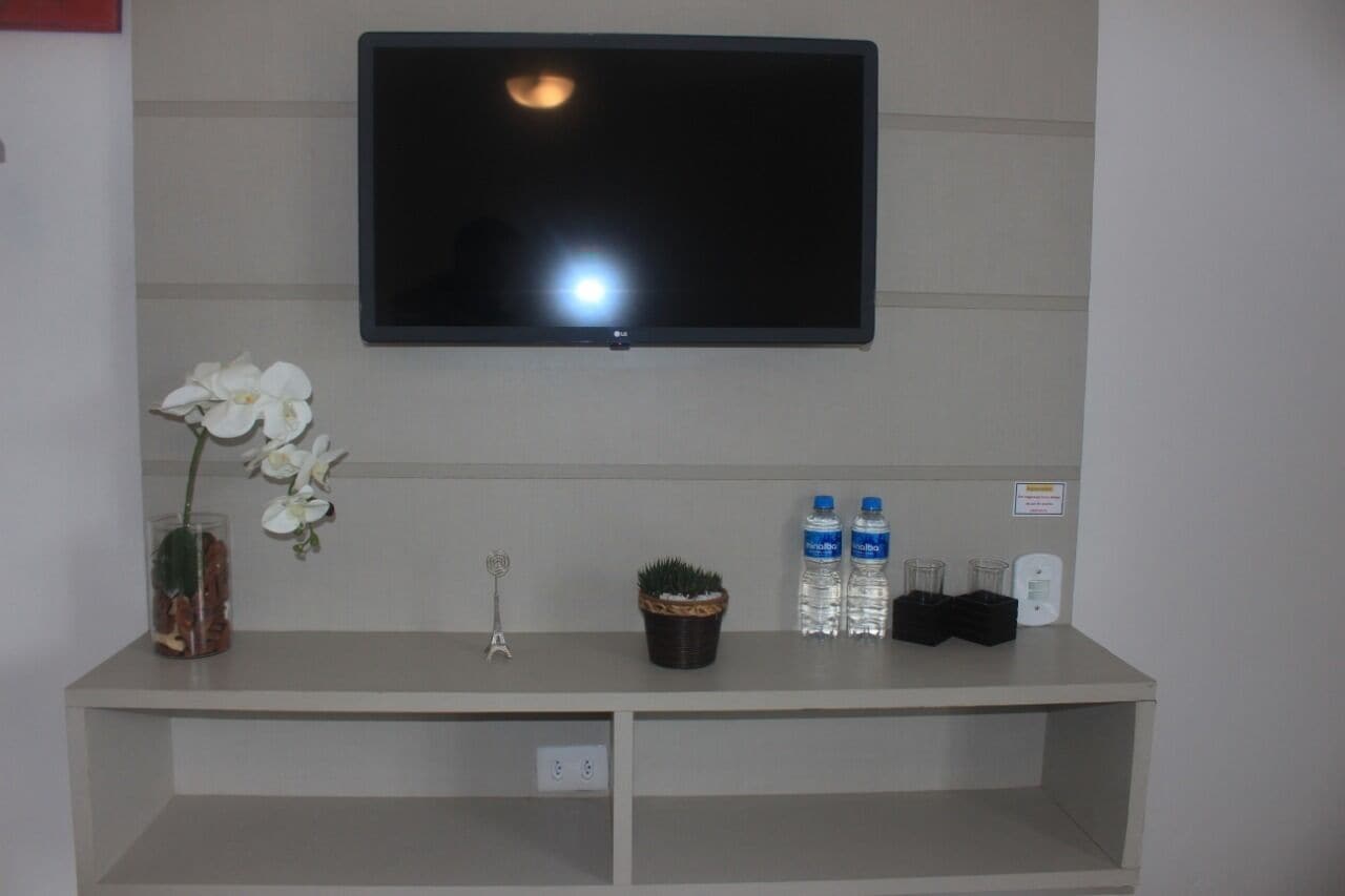 A reception desk with a flat-screen TV, a vase with white flowers, a small potted plant, two water bottles, and a glass set.