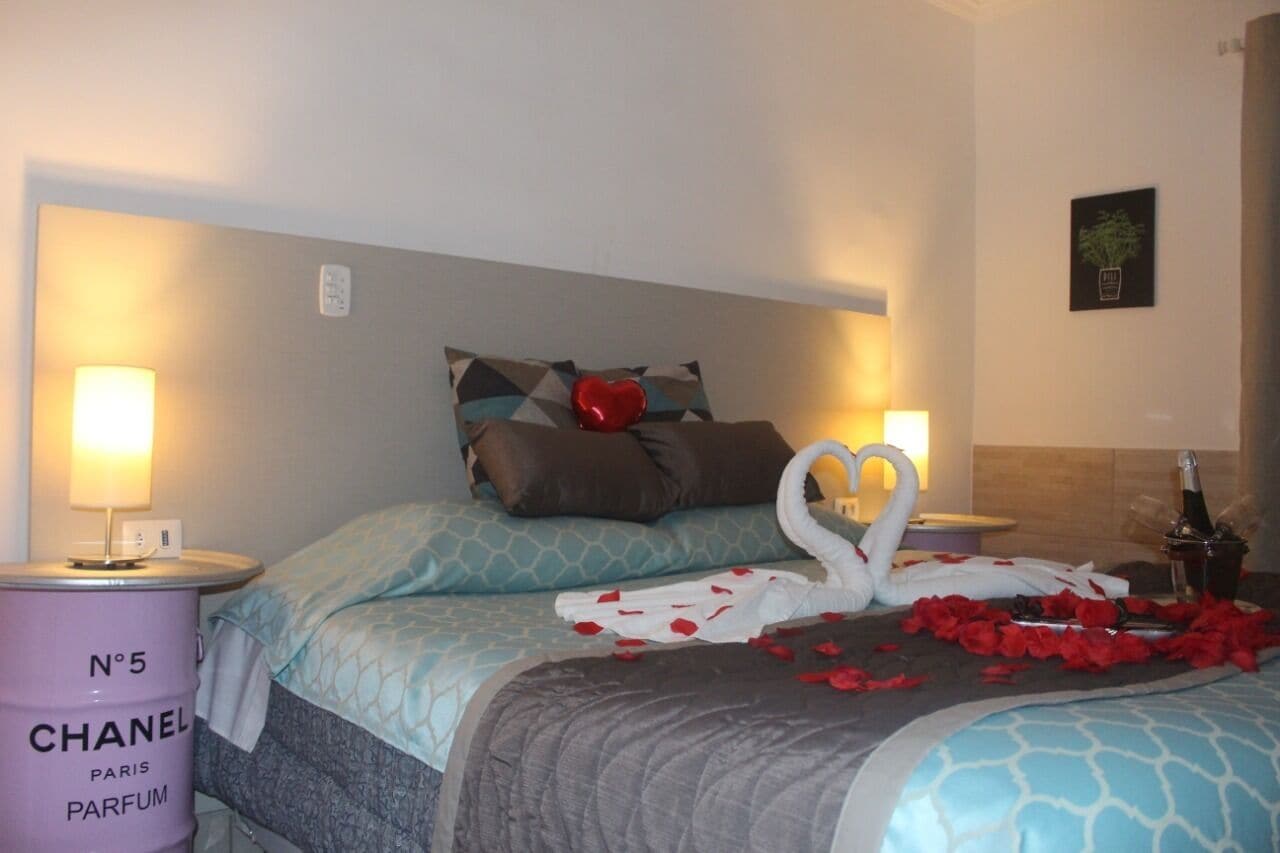 A hotel room with a bed, two bedside lamps, a decorative swan, rose petals, and a bottle of champagne.