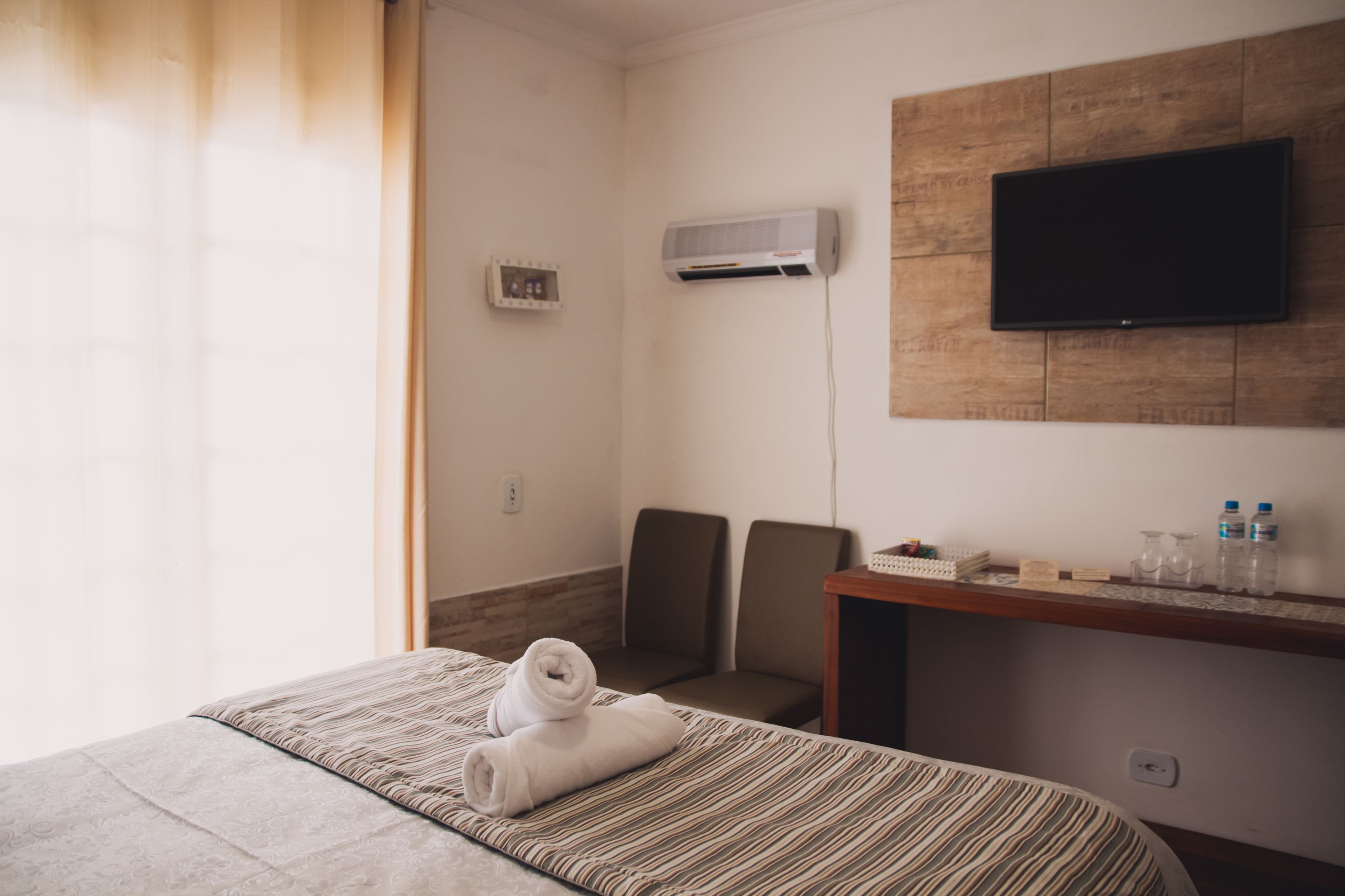 A hotel room with a bed, a desk, a chair, an air conditioning unit, a television, and a window with curtains.