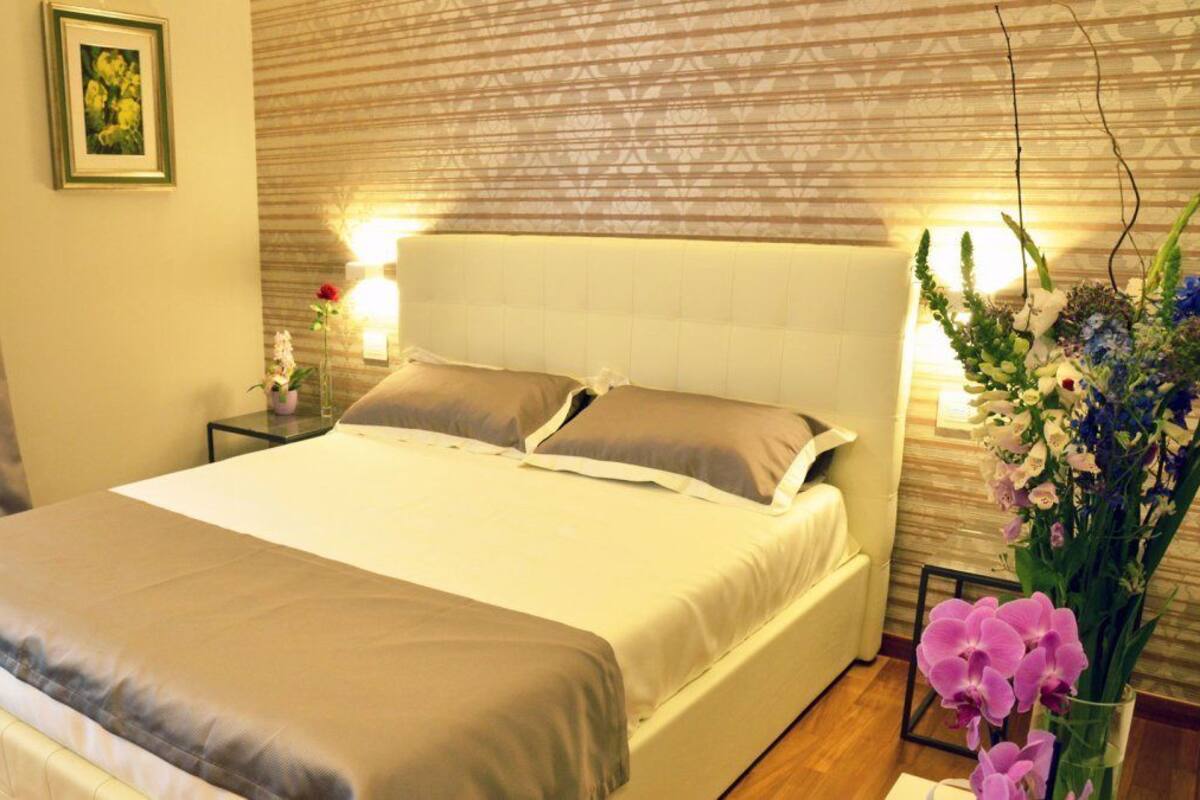A hotel room with a large bed, a nightstand with a lamp, and a floral arrangement.