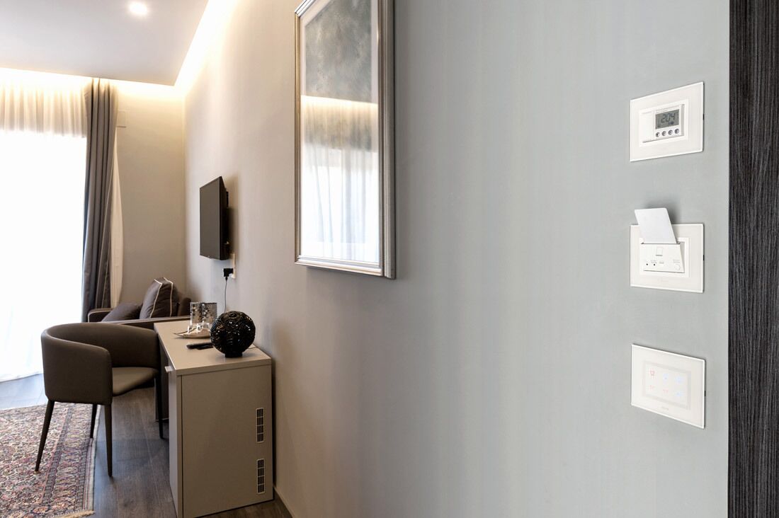 Ein Hotelzimmer mit Schreibtisch, Stuhl und einem großen, gerahmten Bild an der Wand. Drei Lichtschalter und ein Thermostat sind an der Wand befestigt.