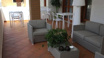 A terrace with grey sofas, a white table, and chairs. There is a potted plant and a lamp on the table.