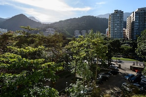 A parking area with cars, surrounded by lush greenery and buildings in the background. - Omar do Rio - Apartamento PUC (Rio de Janeiro)