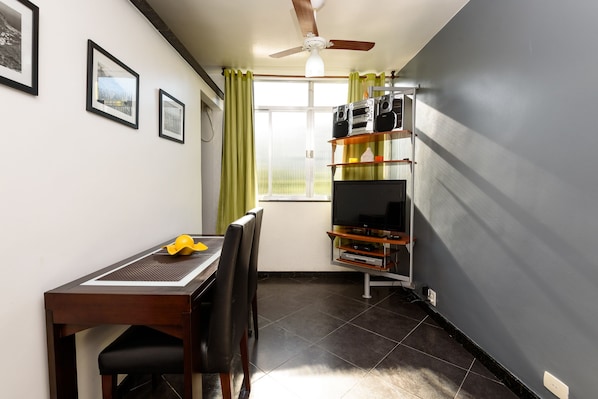 A compact room with a dining table, chairs, a ceiling fan, a TV on a shelf, and framed pictures on the wall. - Omar do Rio - Apartamento PUC (Rio de Janeiro)