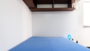 Una habitación con un colchón azul, una manta a rayas de colores, un estante marrón y una lámpara azul.