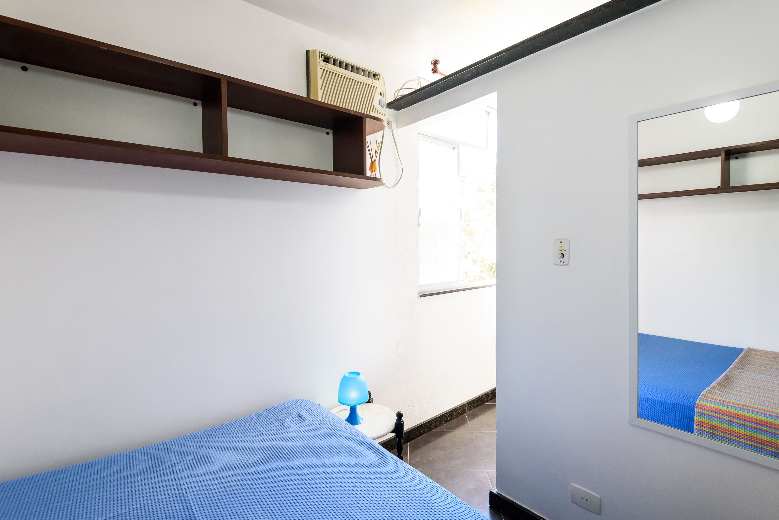 A bedroom with a bed, a nightstand, a lamp, a shelf with a radio, and a window.