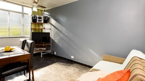 Una sala de estar con un sofá, una mesa de comedor con sillas, un mueble de televisión con dispositivos electrónicos y un ventanal con cortinas.