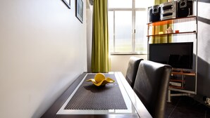 Un comedor con una mesa, sillas, un objeto decorativo amarillo, una ventana con cortinas y una estantería con dispositivos electrónicos.