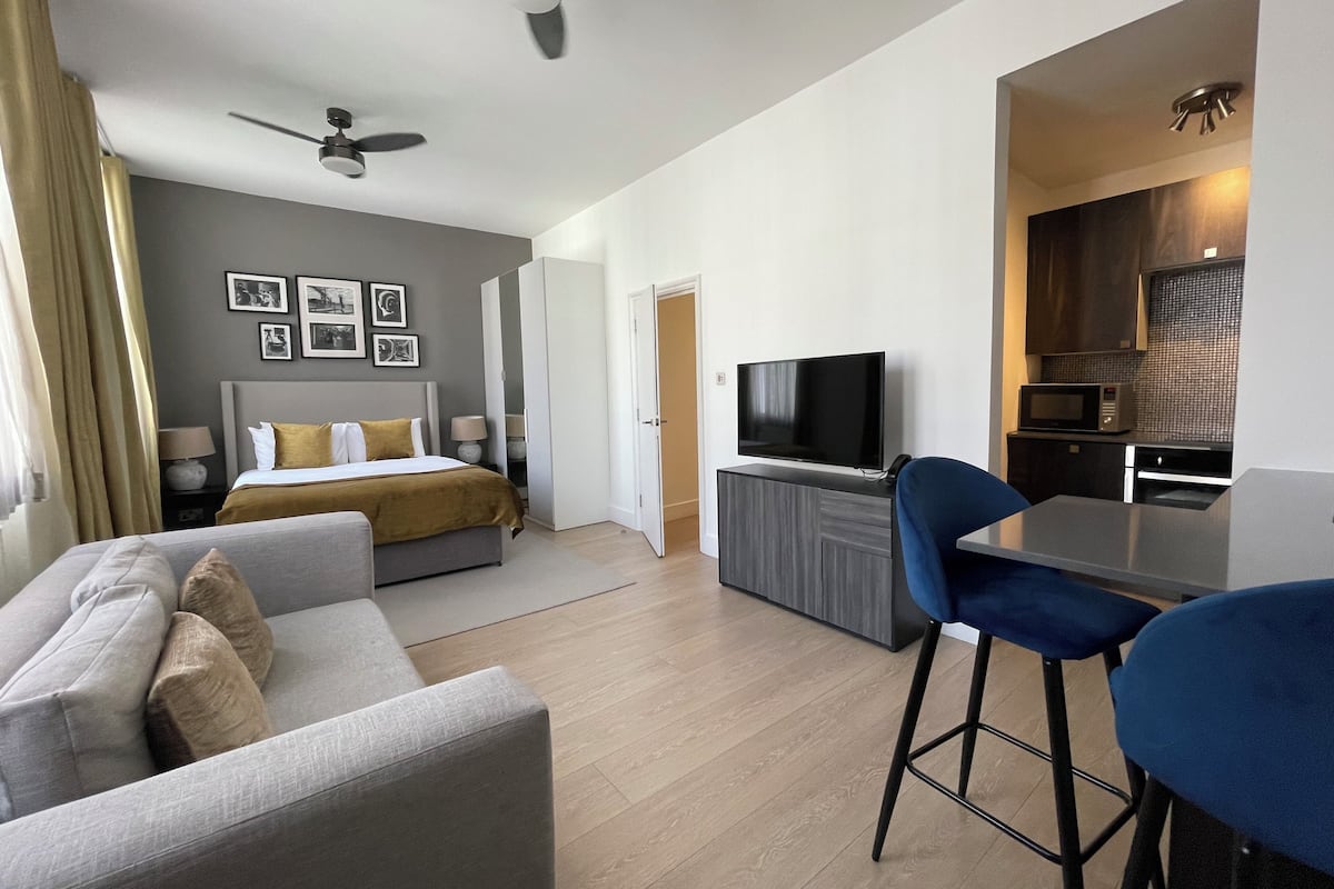 Una habitación de hotel moderna con cama, sofá, televisión, zona de cocina y ventilador de techo.