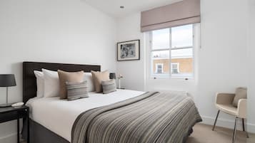 A bedroom with a large bed, a bedside table, a chair, a window with a view of buildings, and a framed picture on the wall.