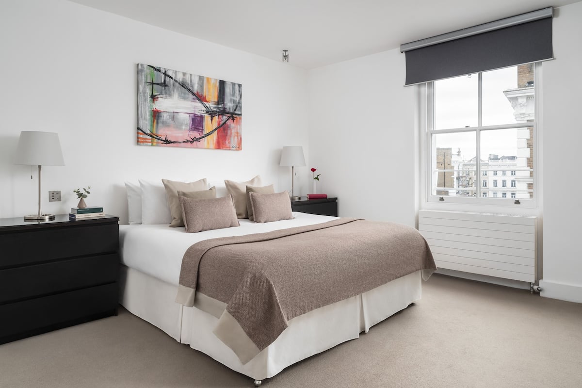 Un dormitorio moderno con una cama grande, una mesita de noche, una lámpara, una ventana con vistas a edificios y un cuadro en la pared.