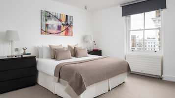 A modern bedroom with a large bed, a nightstand, a lamp, a window with a view of buildings, and a painting on the wall.