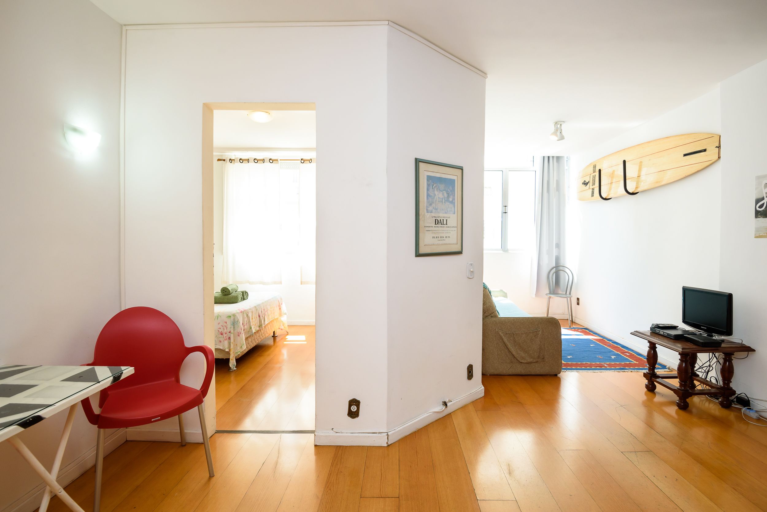A room with a wooden floor, a red chair, a table, a framed picture, a surfboard, and a TV.