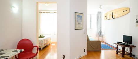 A room with a wooden floor, a red chair, a table, a framed picture, a surfboard, and a TV.