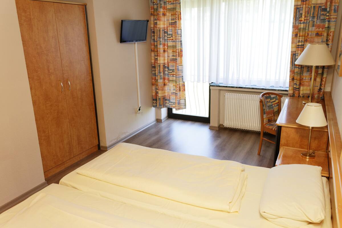 Une chambre d’hôtel avec un lit, un bureau avec une lampe, une chaise, un téléviseur et une fenêtre avec des rideaux.