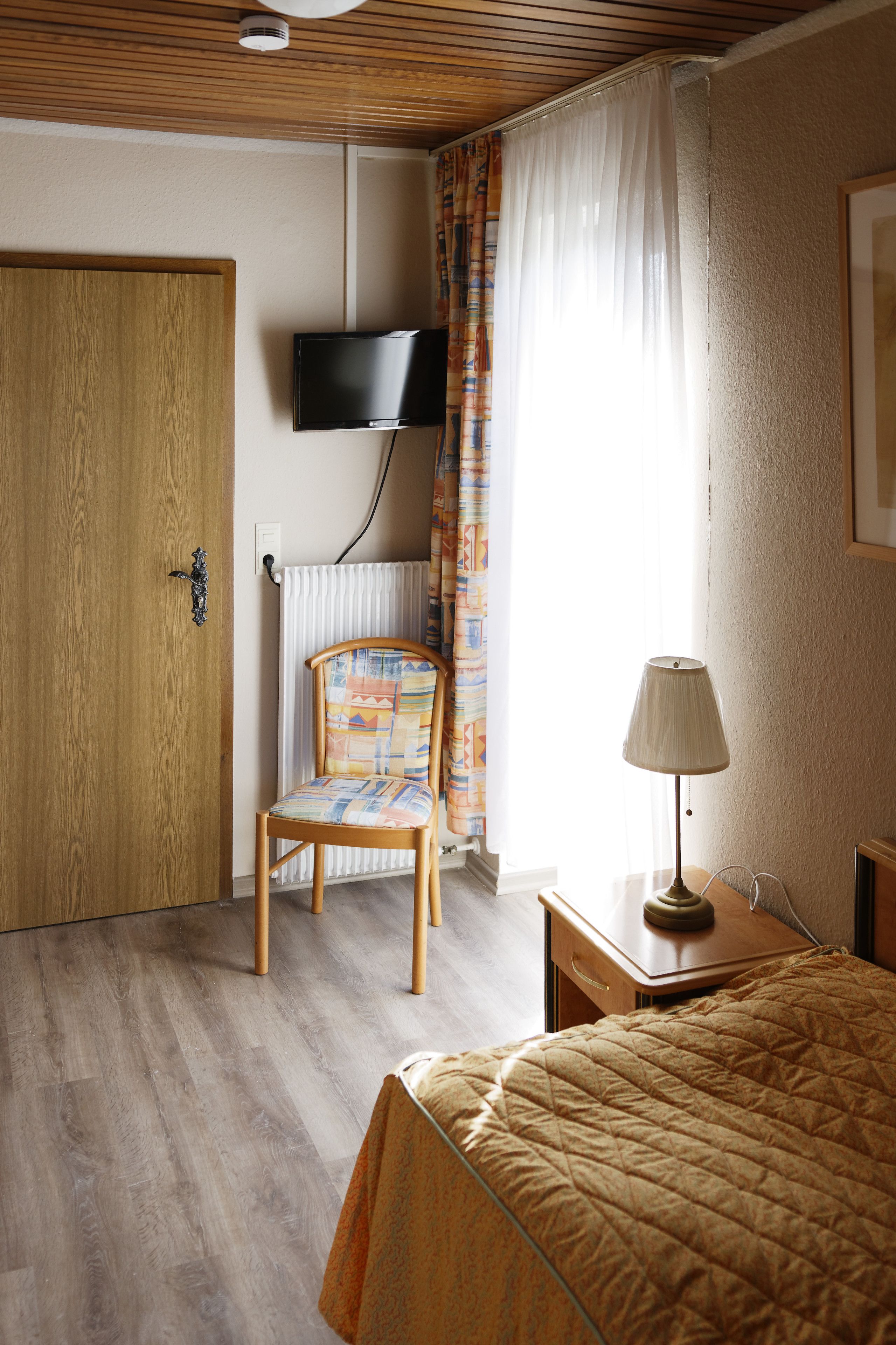A hotel room with a wooden door, a chair, a bed with a quilt, a bedside table with a lamp, a TV mounted on the wall, and a window with curtains.